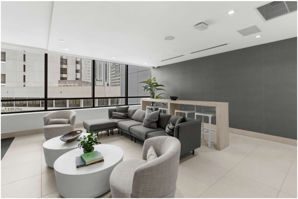 Modern office lounge with large windows, gray sectional sofa, round white coffee tables, armchairs, a tall wooden table with stools, plants, and city buildings visible outside. Neutral color palette and minimal decor.