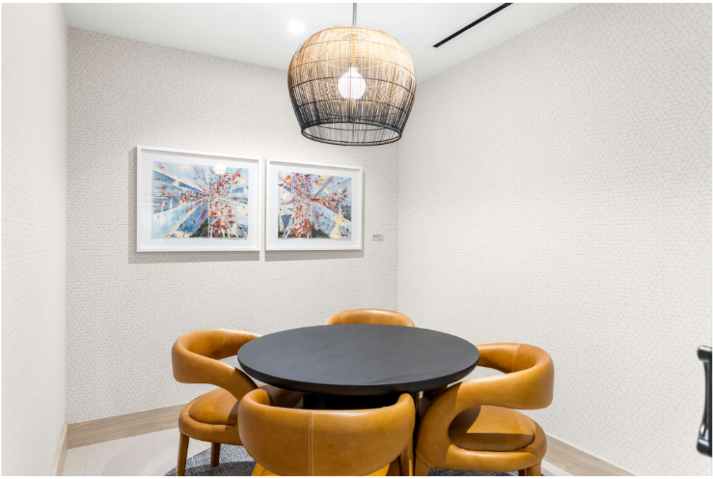 A modern, minimalist dining area with a round black table surrounded by four tan leather chairs, two abstract art pieces on the wall, and a woven pendant light overhead.