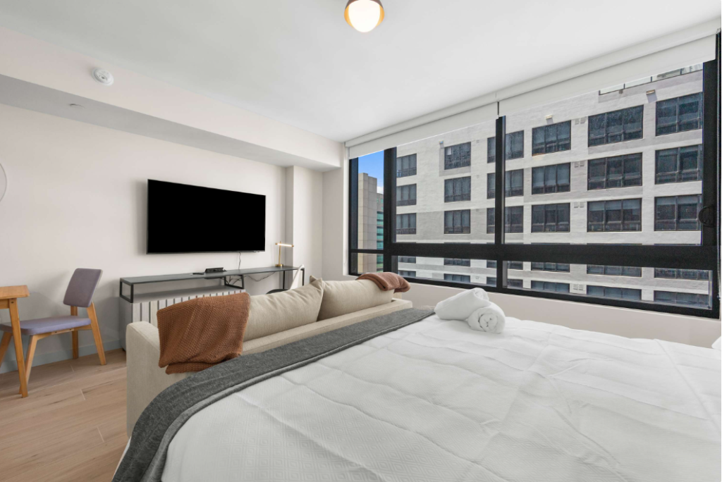 Modern bedroom with a large bed, rolled towels, a sofa, wall-mounted tv, and a desk with a chair. Floor-to-ceiling windows showcase a city view with adjacent tall buildings.