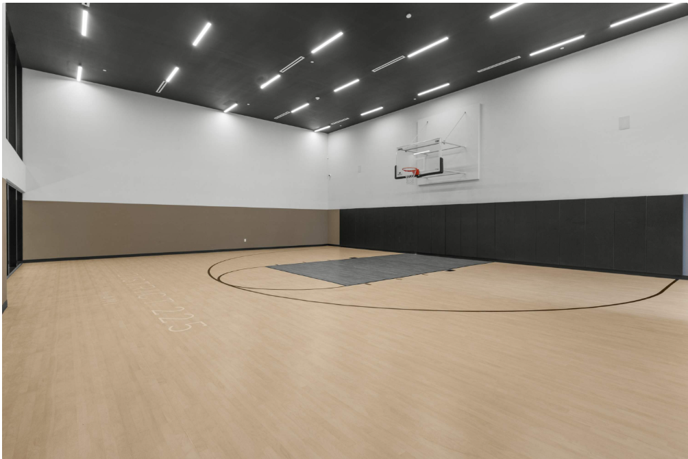 An empty indoor basketball court with light wood flooring, black wall padding, a black-and-white hoop, and bright ceiling lights. The walls are white and tan, and the court is clean and modern.