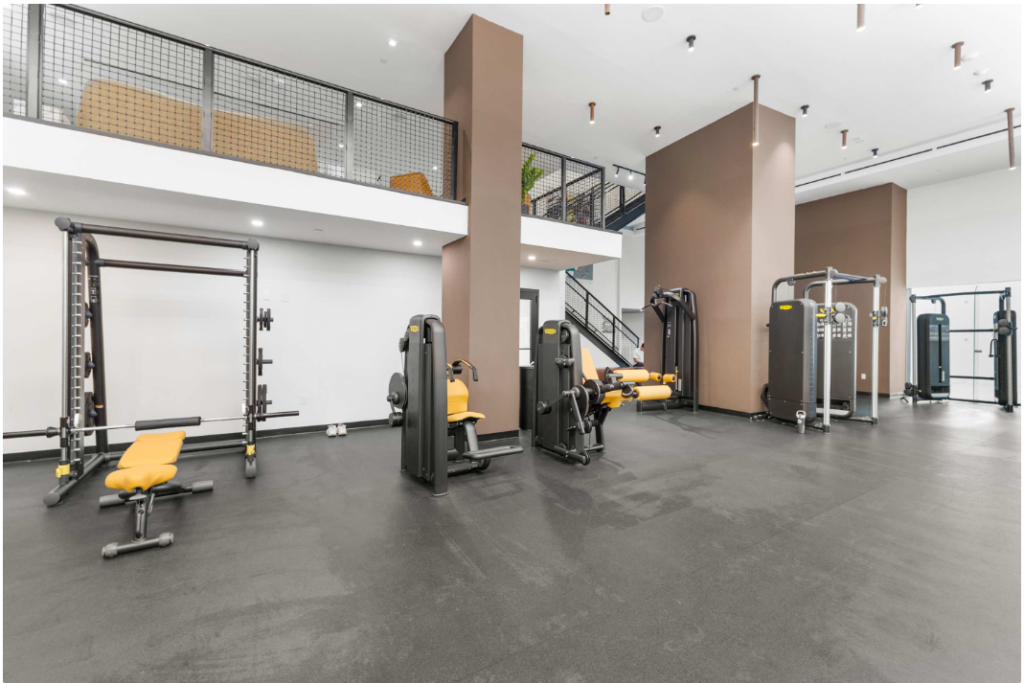 Spacious, modern gym with black and yellow exercise machines, weight racks, and workout benches on a black floor. There is a white wall, brown columns, and a staircase leading to an upper level with a metal railing.