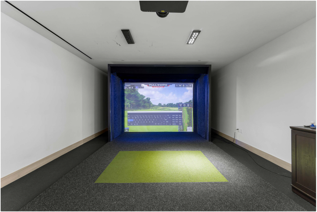 A golf simulator setup in a room, featuring a green mat on the floor and a large screen displaying a golf course and swing data. Overhead lights and a ceiling-mounted projector are visible.