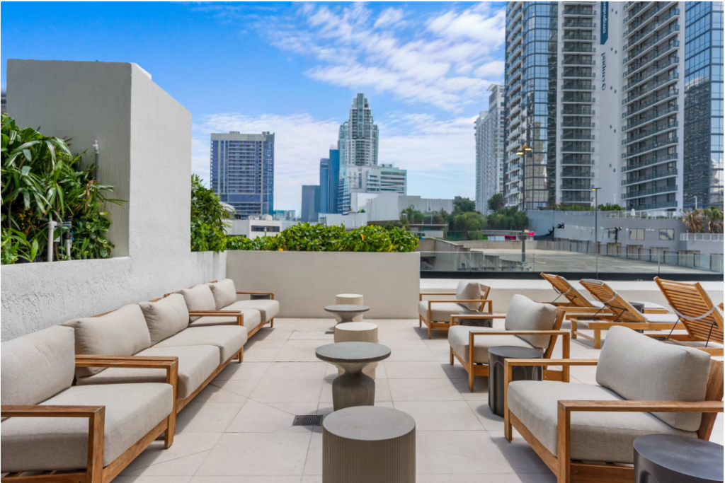 A modern rooftop patio with cushioned wooden chairs, small round tables, and lounge chairs, surrounded by greenery and overlooking tall city buildings under a partly cloudy sky.