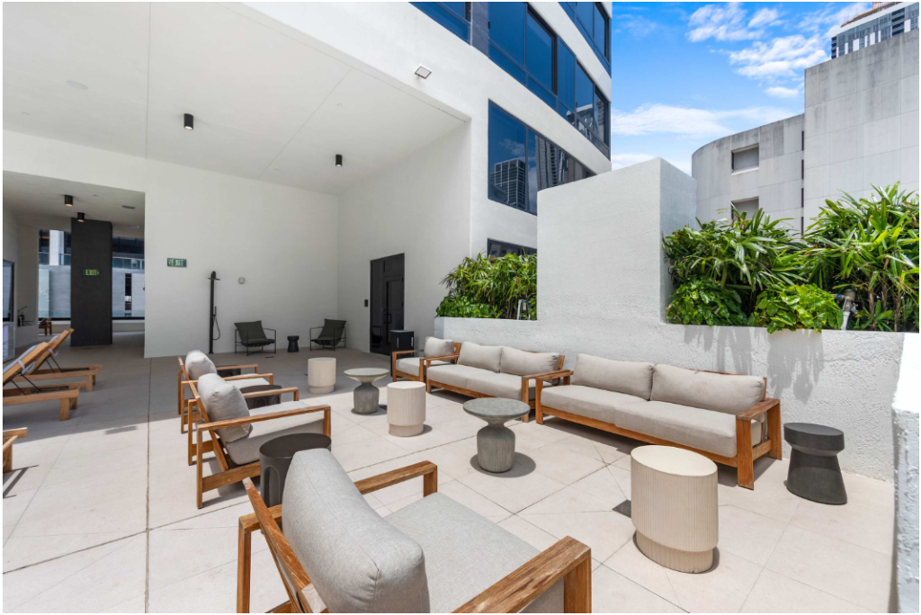 Outdoor lounge area with cushioned wooden chairs, sofas, and small tables arranged on a spacious patio. Green plants line one wall, and tall buildings are visible in the background under a blue sky.
