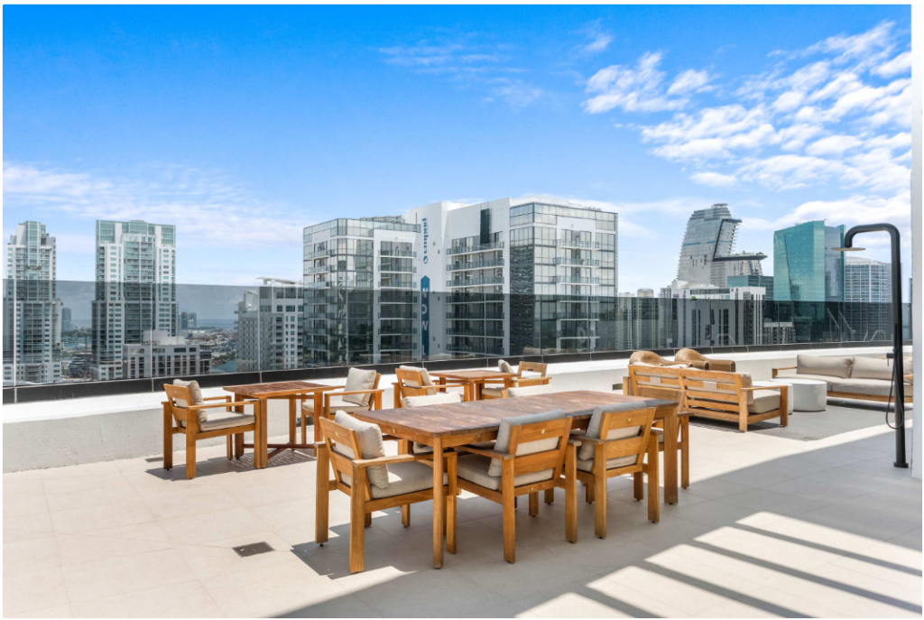 Rooftop patio with wooden tables and chairs, beige sofas, and glass railing overlooking a modern city skyline under a blue sky with scattered clouds.