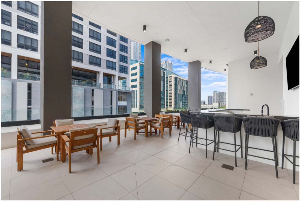 Modern outdoor lounge area with wooden tables and chairs, a bar with woven stools, large columns, and city buildings visible in the background under a partly cloudy sky.