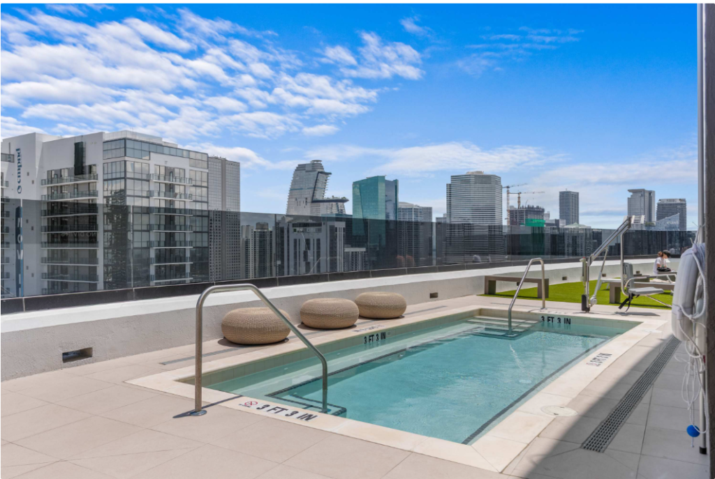 A rooftop pool with handrails, two round loungers, and city skyscrapers in the background under a blue sky with clouds.