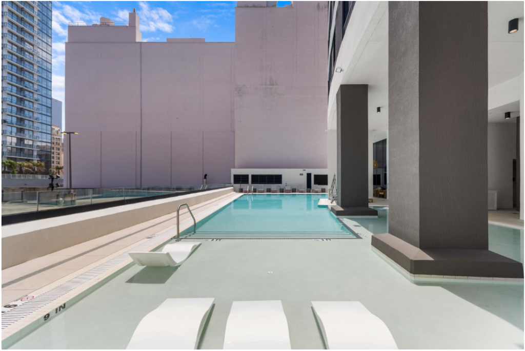 Modern outdoor pool area at an urban high-rise building, featuring shallow water with in-pool lounge chairs, a swimming area, glass safety barriers, and tall city buildings in the background under a bright blue sky.