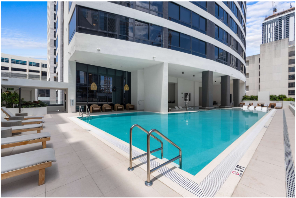 Modern rooftop pool area beside a high-rise building, featuring lounge chairs, a stainless steel pool ladder, and shaded cabanas. Urban skyline and tall buildings are visible in the background under a partly cloudy sky.