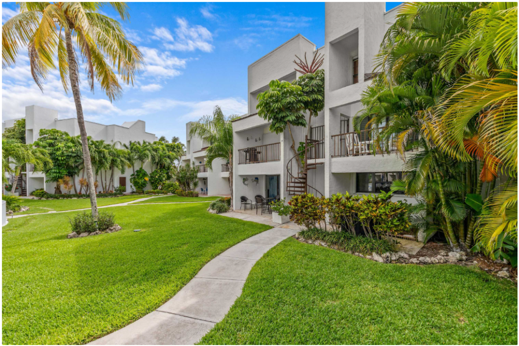 A bright, modern white apartment building with balconies is surrounded by lush green grass, palm trees, and tropical plants under a blue sky with scattered clouds. A curved walkway leads through the landscaped garden.