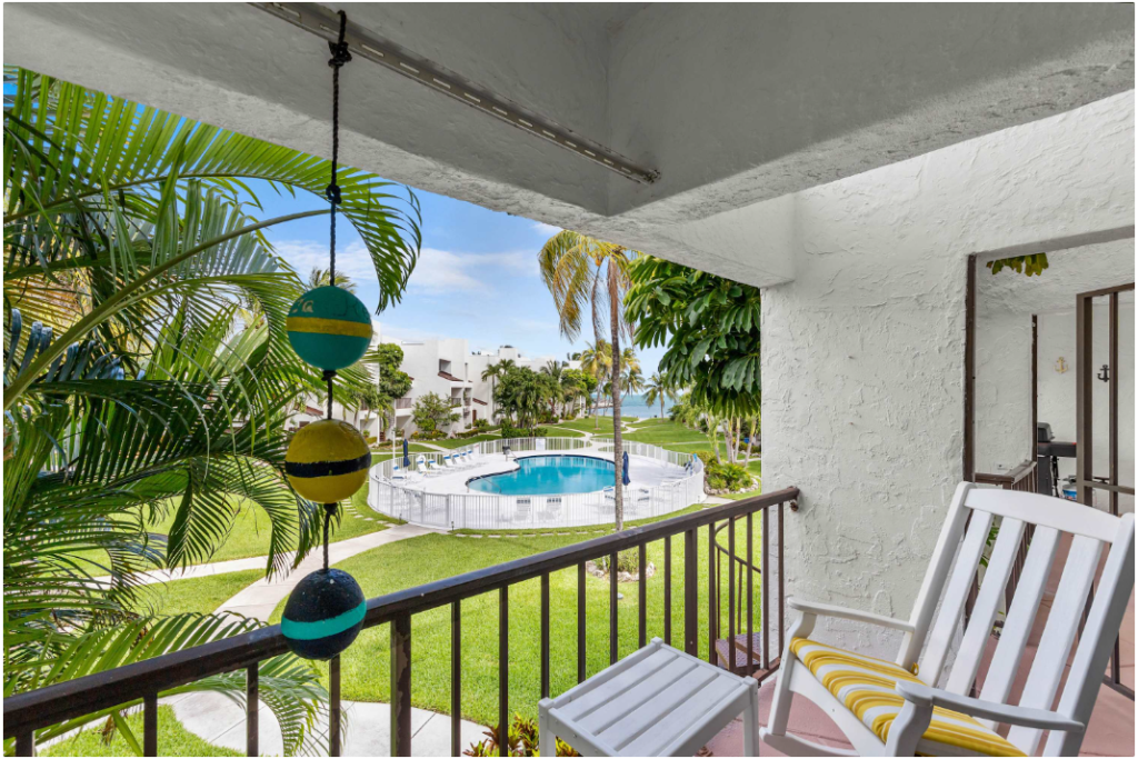 A patio with white rocking chairs overlooks a green lawn, palm trees, a round swimming pool, and white buildings. Three colorful hanging ornaments are in the foreground. A glimpse of water and blue sky is in the background.