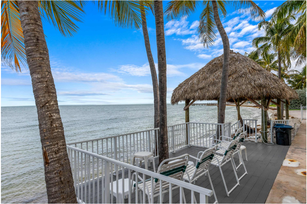 A tropical beachfront scene with palm trees, white lounge chairs on a deck with a white railing, and a thatched-roof tiki hut overlooking calm ocean waters under a blue sky.