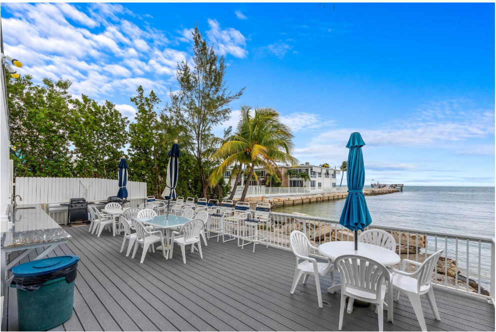 A spacious deck with white plastic chairs and tables, blue umbrellas, and a view of palm trees, a white fence, and the ocean under a partly cloudy sky.