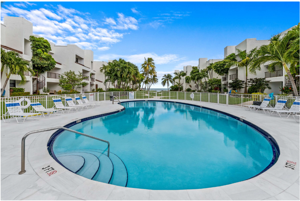 Large, oval outdoor swimming pool surrounded by white lounge chairs and palm trees, with modern white buildings on either side and the ocean visible in the background under a bright blue sky.