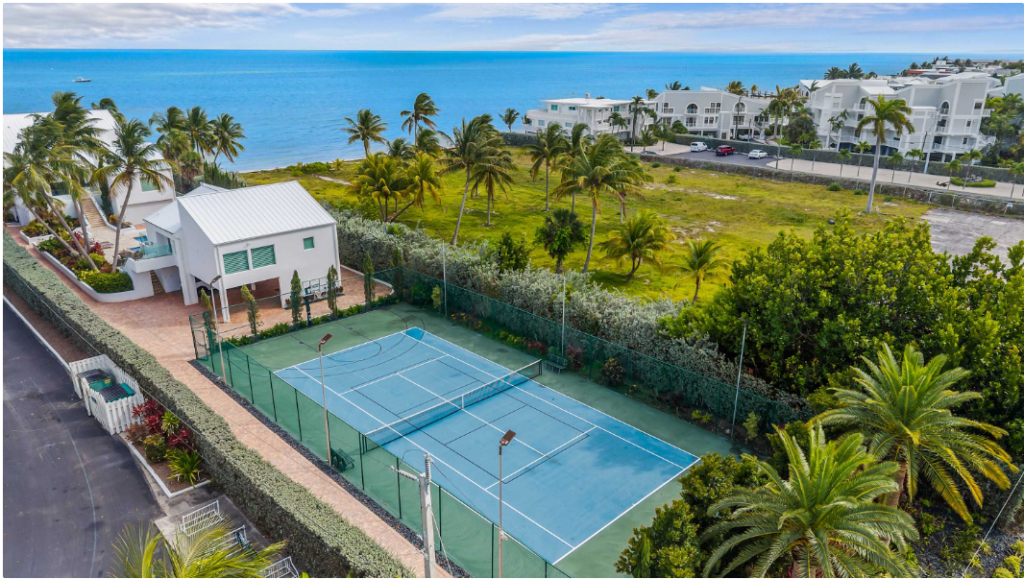 Aerial view of a tennis court surrounded by palm trees near the ocean, with white buildings and green lawns in the background under a partly cloudy sky.