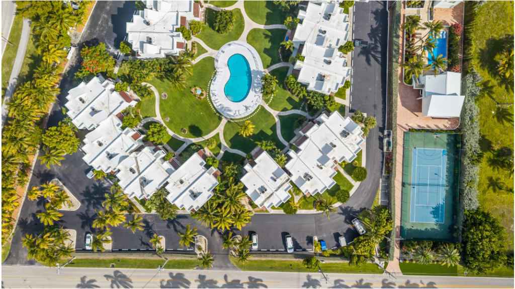 Aerial view of a circular apartment complex with a central swimming pool, surrounded by green lawns, palm trees, parked cars, a tennis court, and nearby roads.