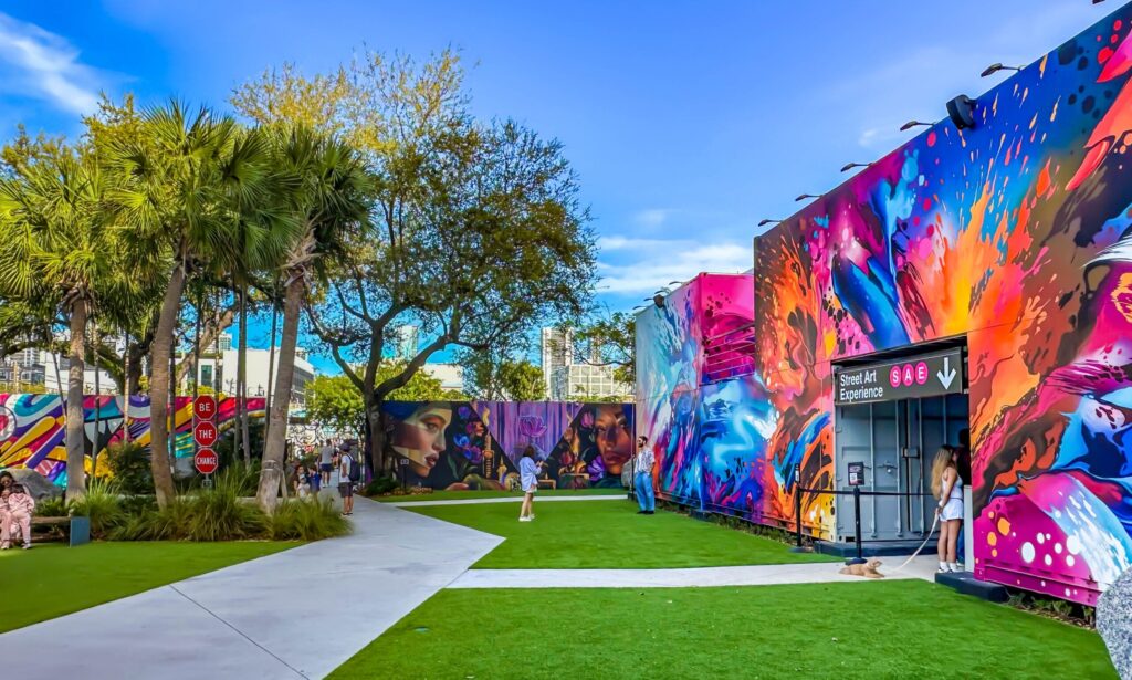 A colorful outdoor art space with vibrant murals covering large walls, green grass, palm trees, and people walking along pathways under a clear blue sky.