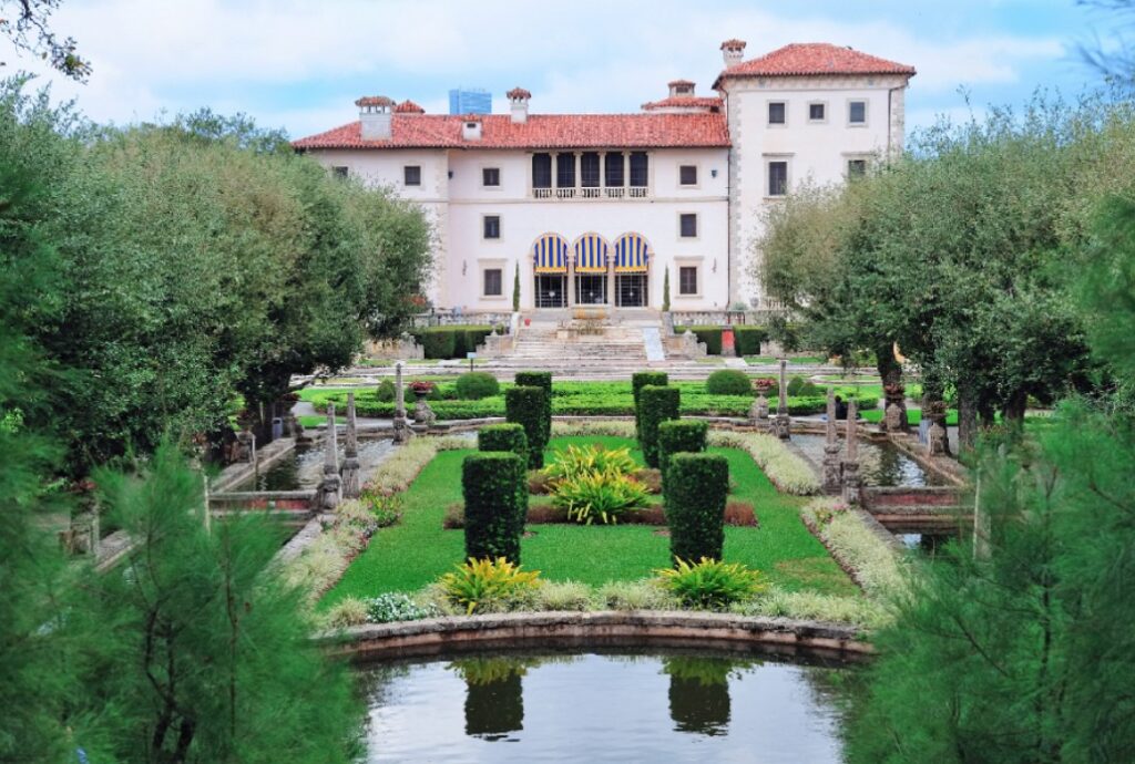 A grand villa with a red-tiled roof stands behind formal gardens featuring manicured hedges, topiary, fountains, and a reflective pond, all framed by lush greenery and trees.