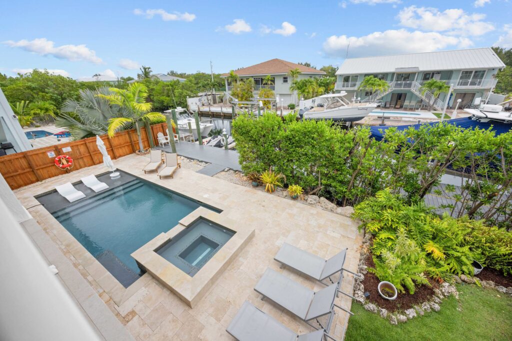 Aerial view of a backyard with a small rectangular pool, attached hot tub, lounge chairs, and lush green landscaping. There is a wooden fence, umbrellas, and a canal with docked boats beyond the yard.