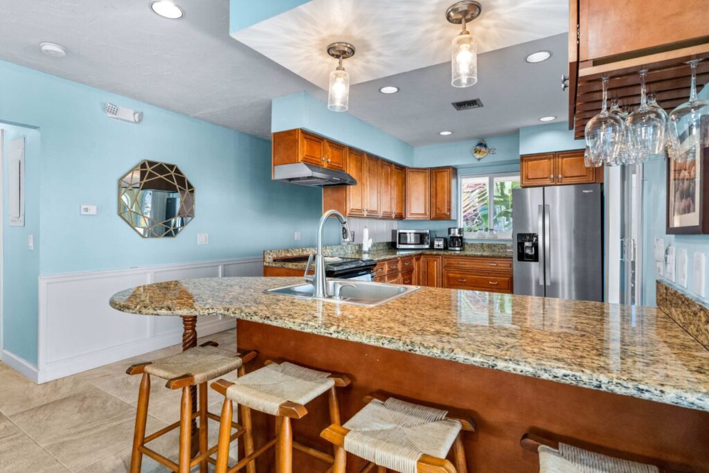 A modern kitchen with wooden cabinets, granite countertops, and stainless steel appliances. Three wooden barstools sit at a breakfast bar under pendant lights, with glasses hanging overhead. Walls are painted light blue.