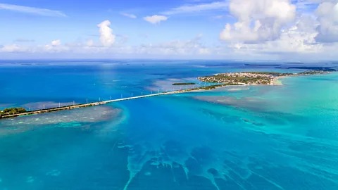 A long bridge stretches across turquoise blue ocean waters, connecting small islands under a bright, partly cloudy sky. The surrounding sea features varying shades of blue and green.
