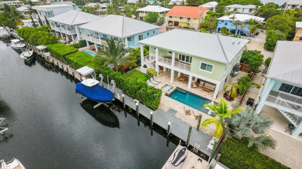 Aerial view of waterfront homes with pools along a canal, docked boats, lush greenery, and palm trees in a residential neighborhood.