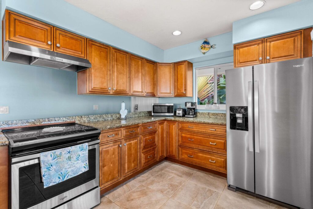 A modern kitchen with wooden cabinets, stainless steel appliances, granite countertops, a stove with a tropical-patterned towel, a refrigerator, microwave, and a window above the sink with a ceramic fish decoration.