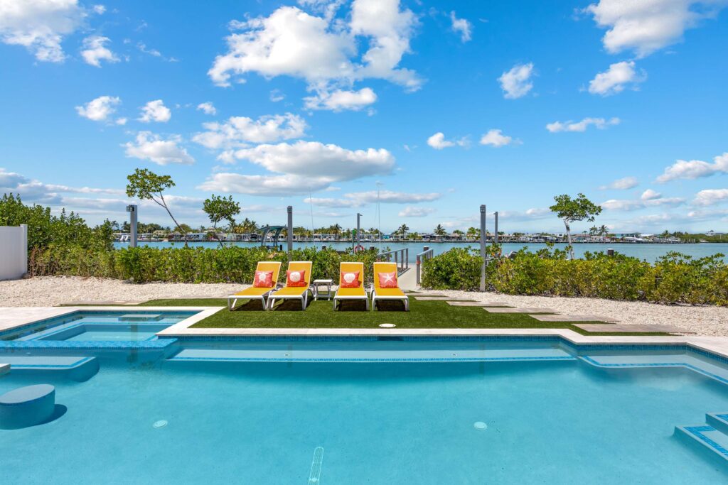 A backyard pool with a hot tub, three yellow lounge chairs on green grass, and a view of water and distant houses under a blue sky with scattered clouds.