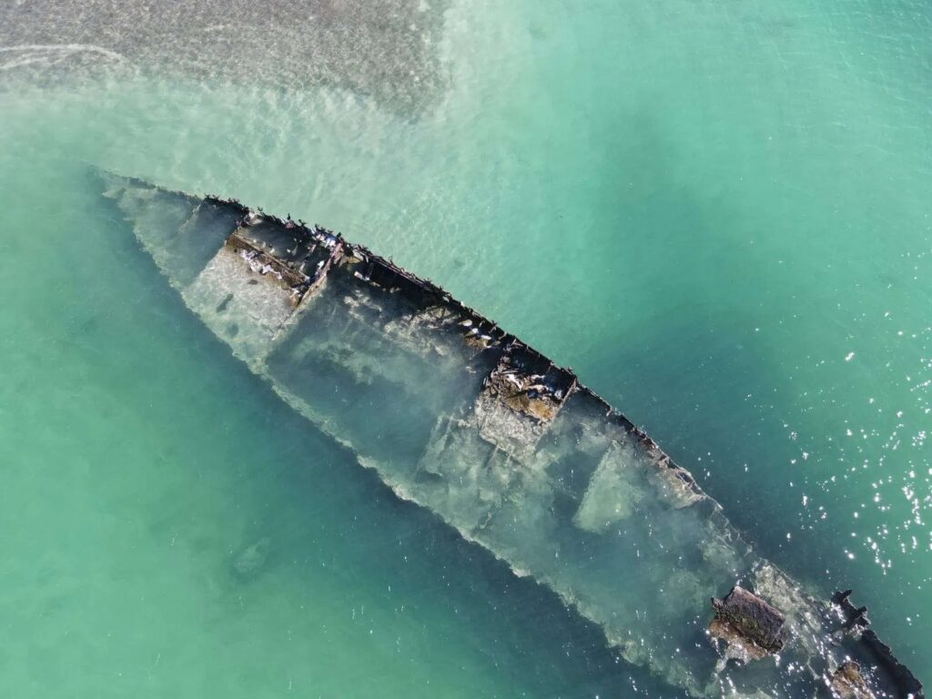 Aerial view of a partially submerged shipwreck in clear turquoise water, with the outline of the rusted vessel visible beneath the surface.