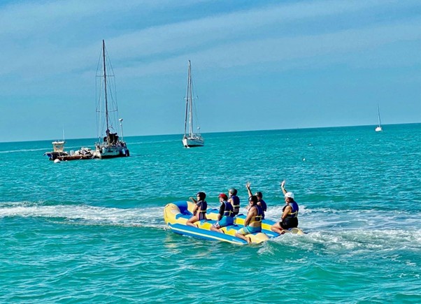 Five people in life jackets ride an inflatable banana boat on turquoise water, with two sailboats and a clear blue sky in the background. Some riders raise their hands, appearing to enjoy the activity.