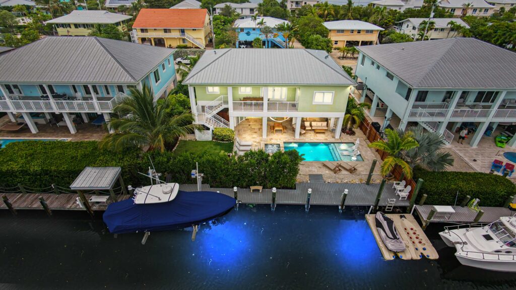 Aerial view of waterfront homes with pools, palm trees, and docks. One dock has a covered boat and blue underwater lights illuminating the water at dusk.
