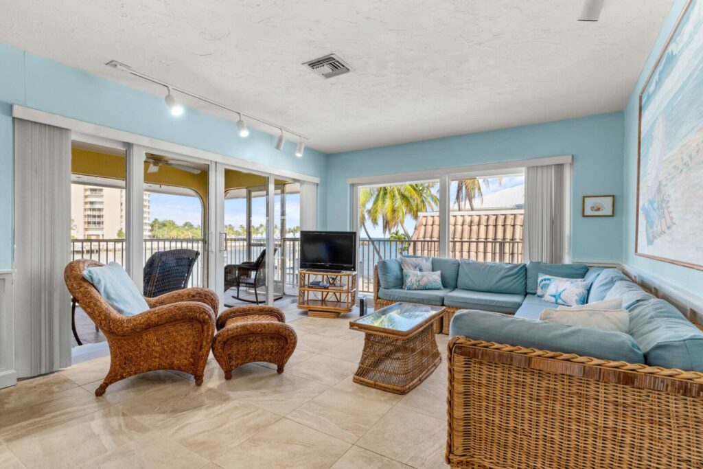 A bright living room with light blue walls, wicker furniture, a glass coffee table, a flat-screen tv, and large windows offering views of palm trees and a covered patio.