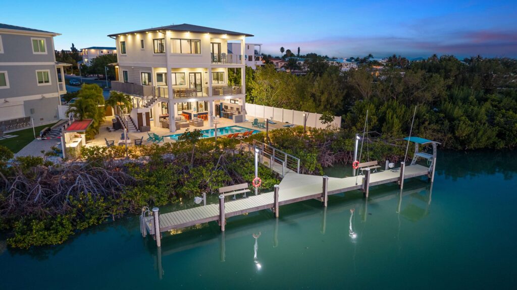 A modern, three-story waterfront house with large windows and balconies, an outdoor pool area, and a private dock extending over calm blue water at dusk, with trees and surrounding houses nearby.