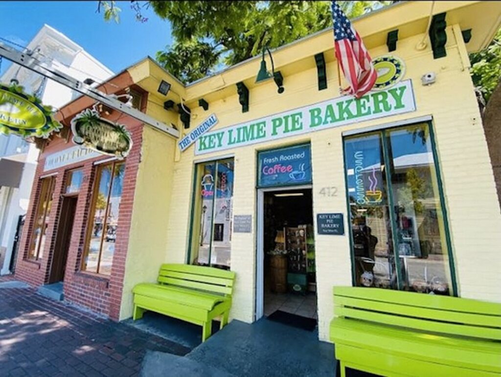A yellow bakery storefront with a sign reading key lime pie bakery, green benches outside, an american flag above, and a brick building next door. Trees and a sunny sky are reflected in the windows.