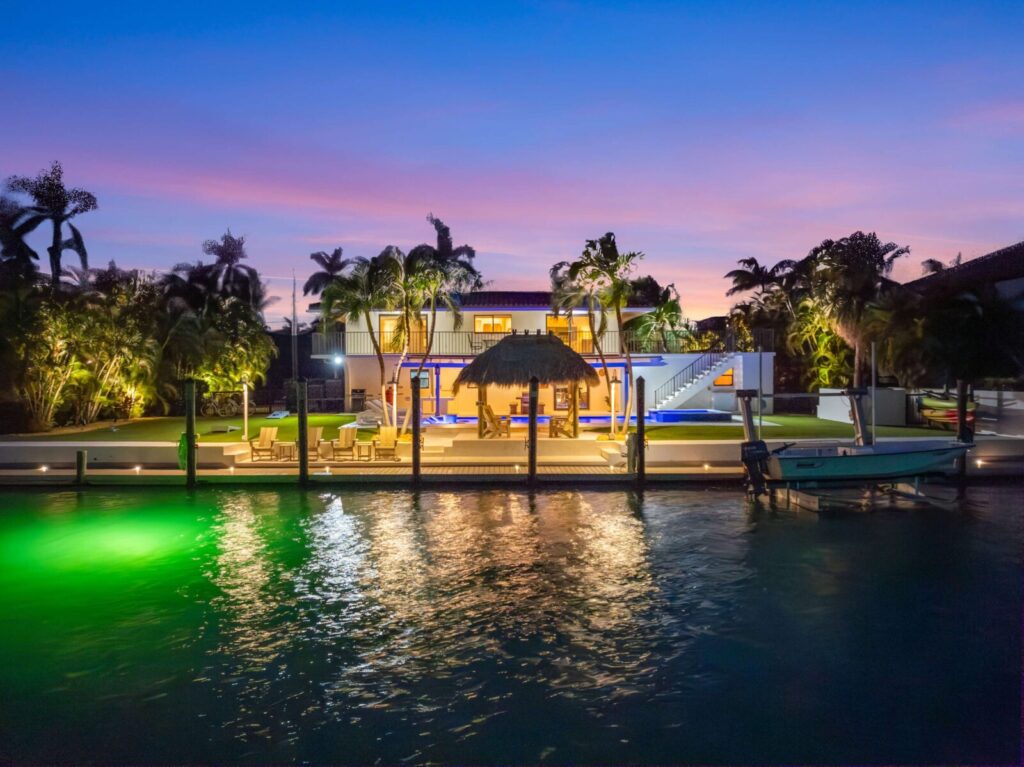 Modern waterfront house illuminated at dusk, surrounded by palm trees, with a thatched-roof patio, outdoor seating area, stairs, and a docked boat on calm water glowing with green underwater lights.