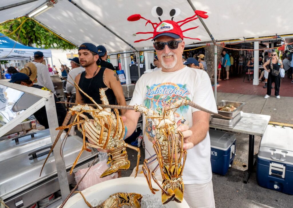 A smiling man wearing a crab hat and sunglasses holds two large lobsters at an outdoor seafood festival, with other people and coolers visible under a tent in the background.