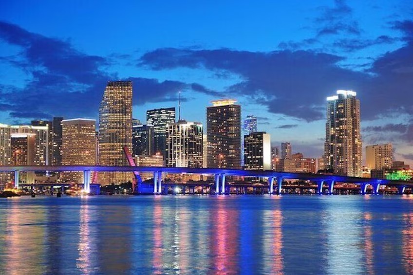Miami city skyline at dusk, with illuminated skyscrapers reflecting on the water and a bridge lit in blue crossing the foreground under a partly cloudy evening sky.