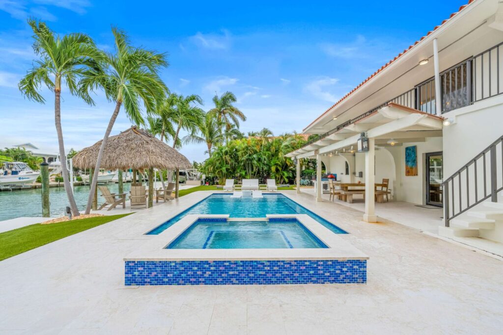 Elegant backyard with a pool and spa, palm trees, a thatched-roof tiki hut, lounge chairs, and a covered patio dining area beside a white waterfront house. Blue sky and boats are visible in the background.