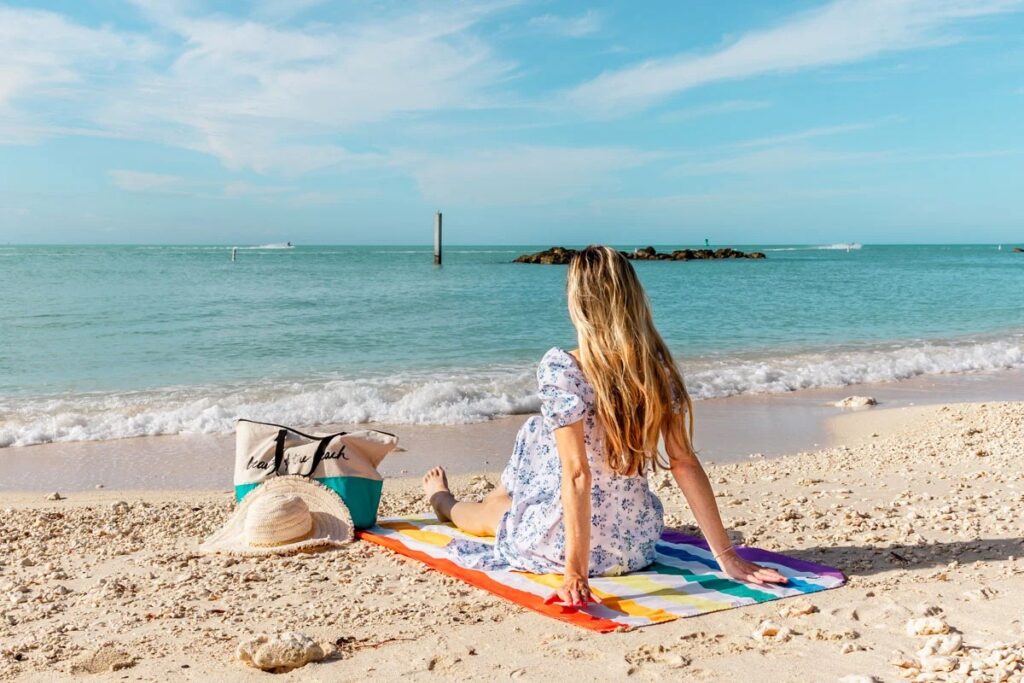 What to pack for florida keys: the only checklist (2026) 2 A woman in a light blue floral dress sits on a colorful striped towel at a sandy beach, facing the sea. A tote bag and a sun hat are next to her, with clear blue sky and calm water in the background.