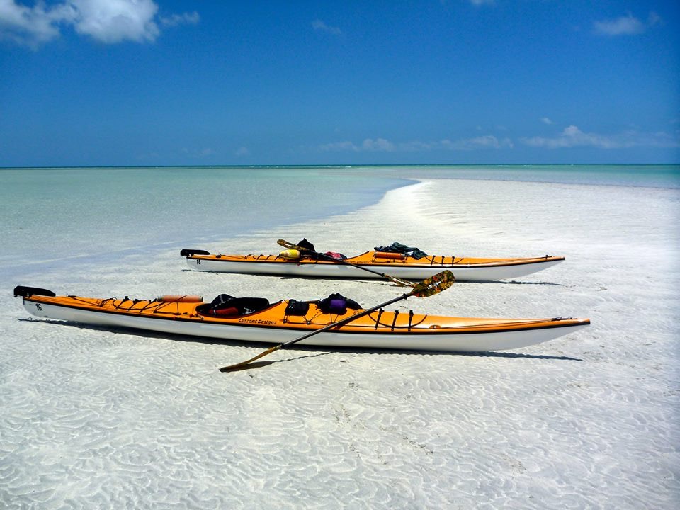 Two yellow kayaks rest on a shallow, sandy beach with clear turquoise water under a bright blue sky, suggesting a peaceful tropical setting.