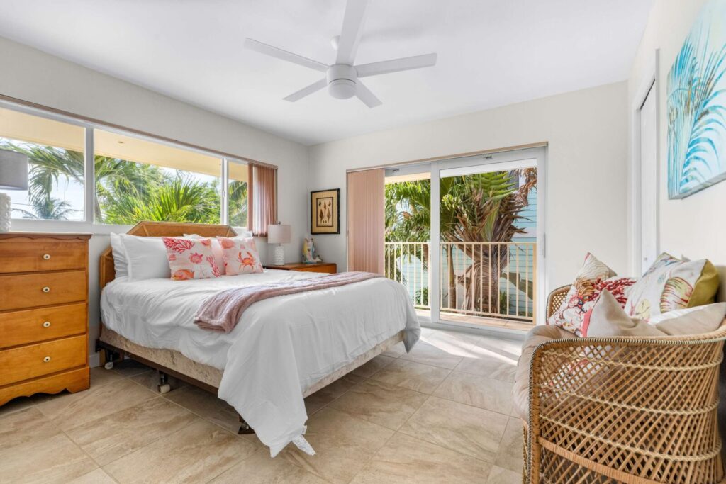 A bright bedroom with a large bed, wicker chair, dresser, and windows showing palm trees outside. Light tones and beach-themed decor create a relaxed, airy atmosphere.
