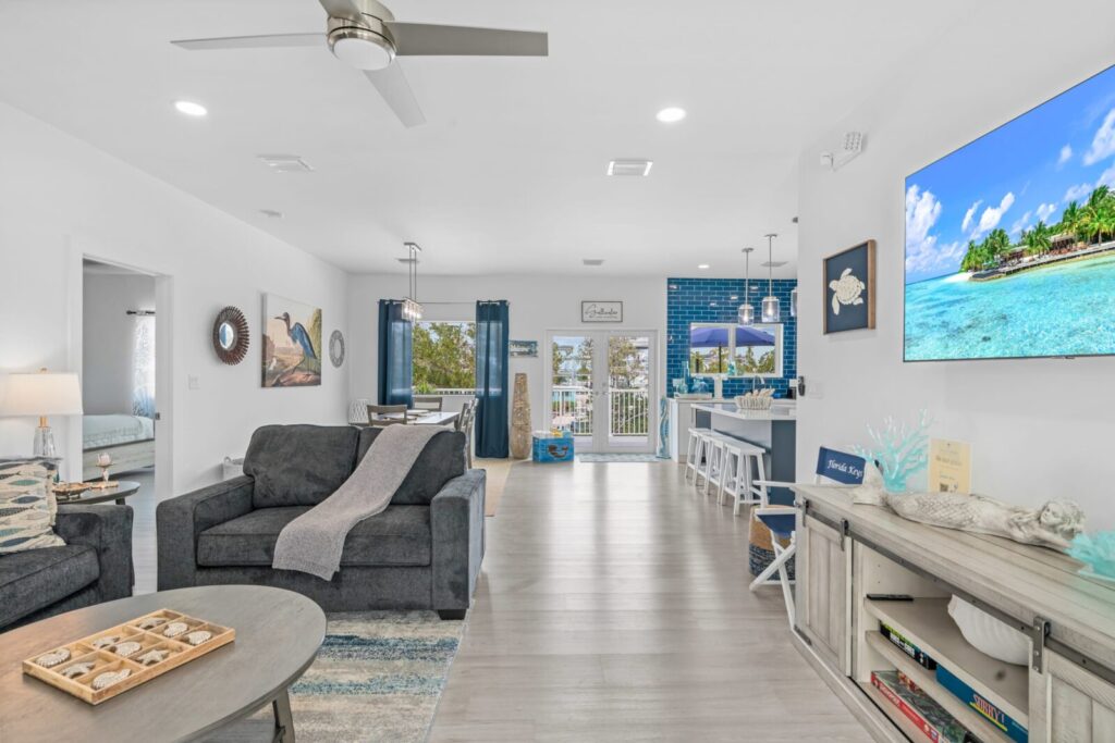 Open-concept living space with gray sofas, coastal decor, wall-mounted tv, and a blue-tiled kitchen. Large windows and glass doors offer natural light and a view of a patio and greenery outside.