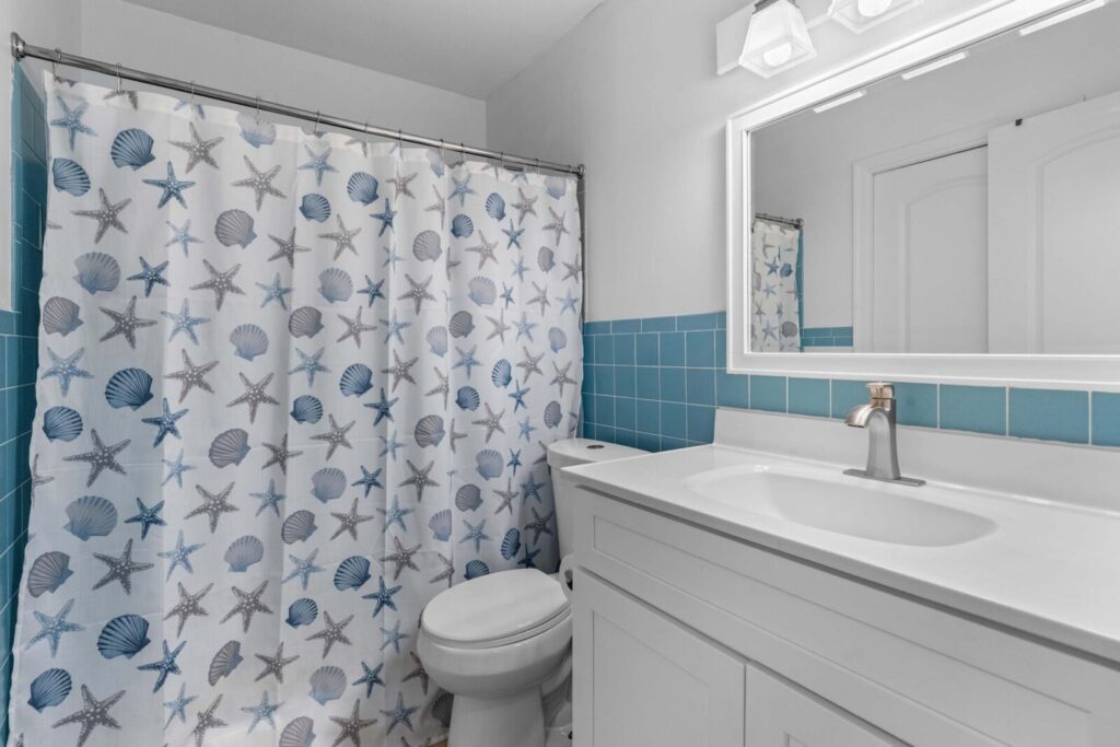 A bathroom with a white vanity, sink, and mirror, blue-tiled walls, and a shower curtain decorated with seashell and starfish patterns in blue and gray tones. Toilet is next to the sink.