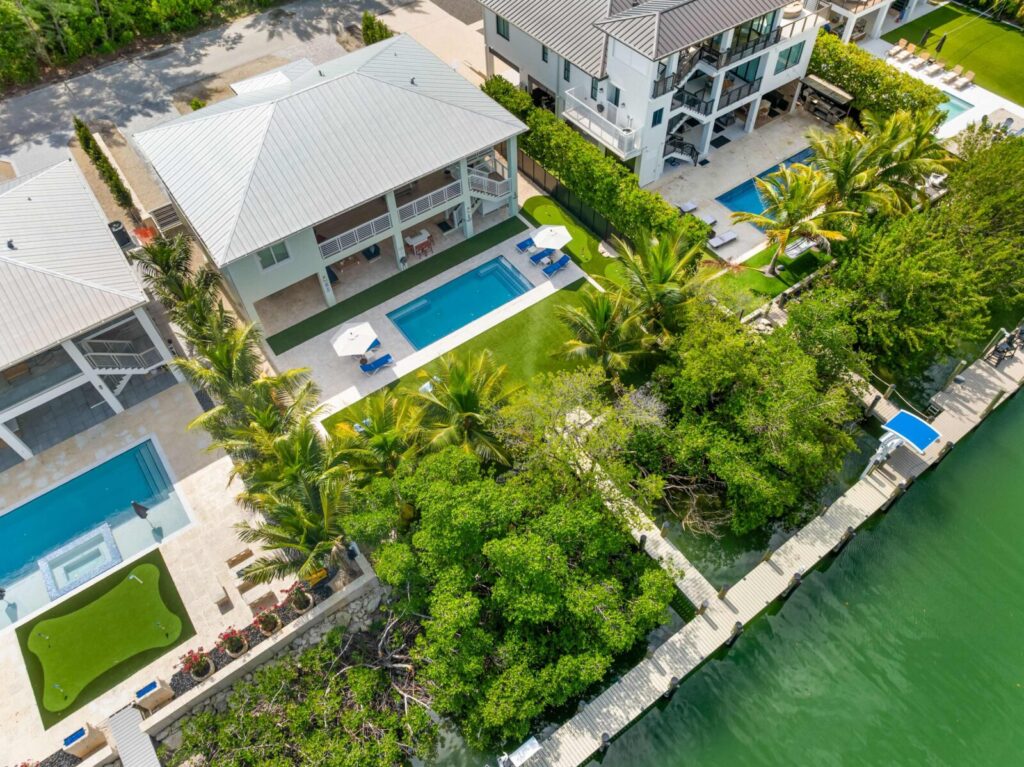 Aerial view of modern waterfront homes with pools, palm trees, a putting green, and a dock along a calm green canal. Lush greenery borders the properties, and outdoor seating areas are visible.