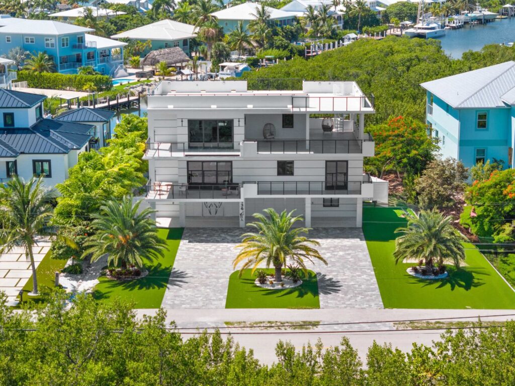 What to pack for florida keys: the only checklist (2026) 7 A modern three-story white house with large windows, surrounded by lush greenery, palm trees, and a stone driveway, located in a coastal neighborhood with water and other colorful houses nearby.