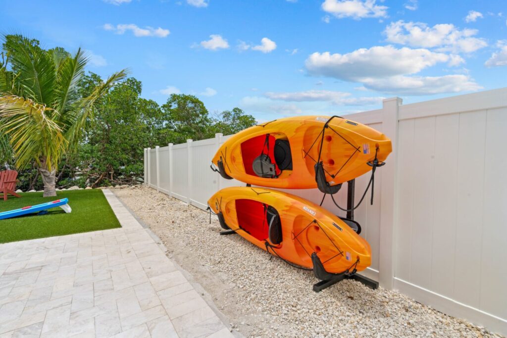 What to pack for florida keys: the only checklist (2026) 5 Two bright orange kayaks are stacked on a white fence in a backyard with stone pavement, artificial grass, palm tree, and a blue sky with scattered clouds.