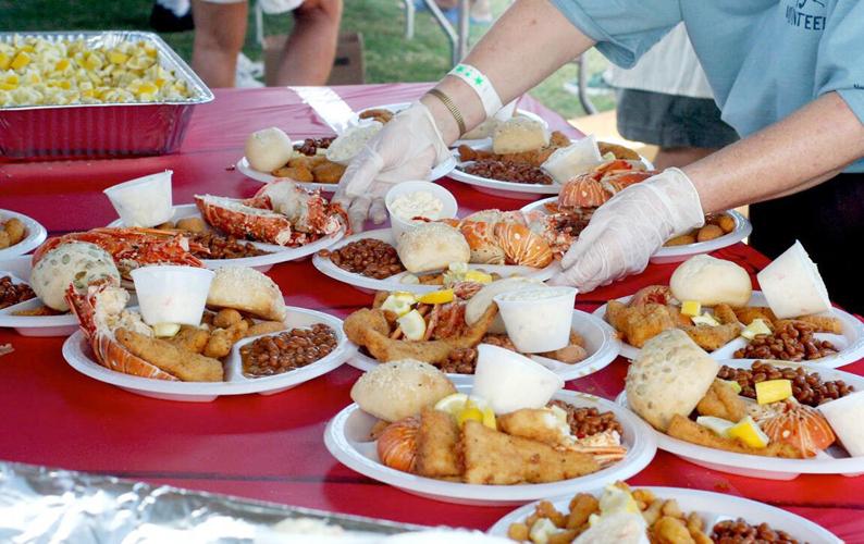 A person wearing gloves serves plates of food on a red table, each plate containing lobster, corn, baked beans, a roll, and a cup of coleslaw at an outdoor event. Other people are visible in the background.