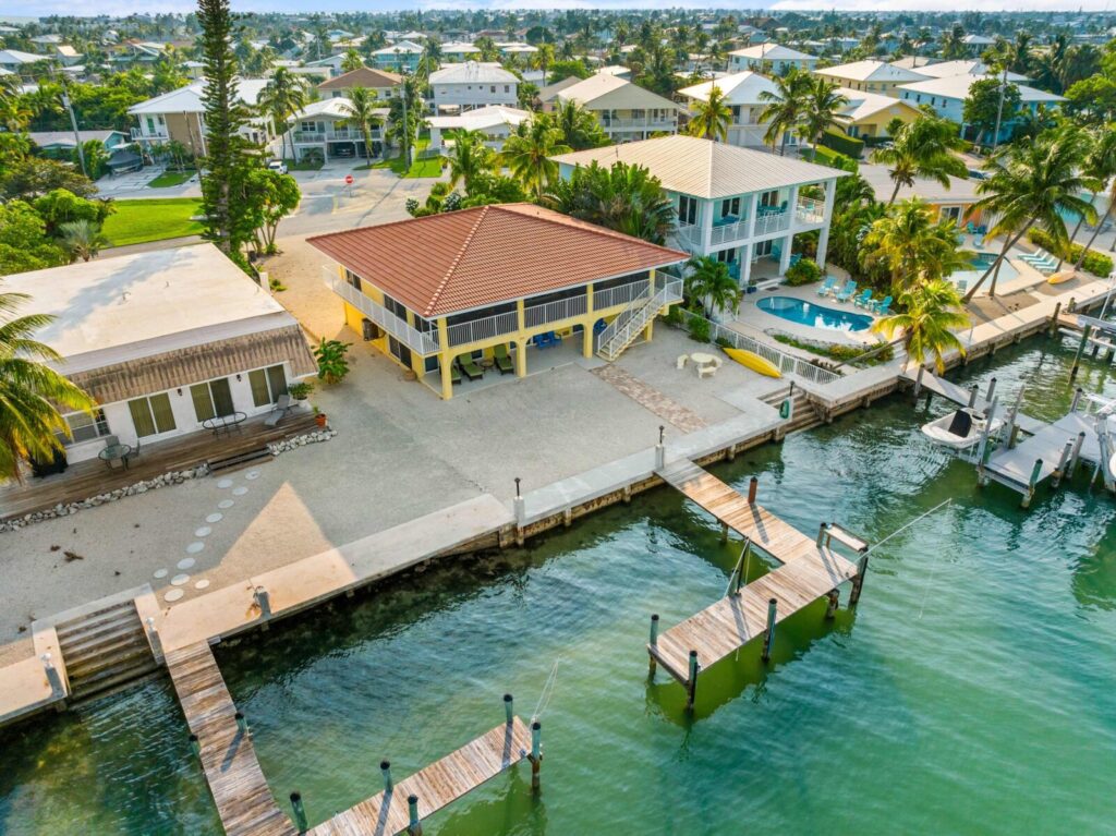 Aerial view of waterfront homes with docks along a canal, featuring a two-story house with a red roof and yellow pillars, surrounded by palm trees and a swimming pool.