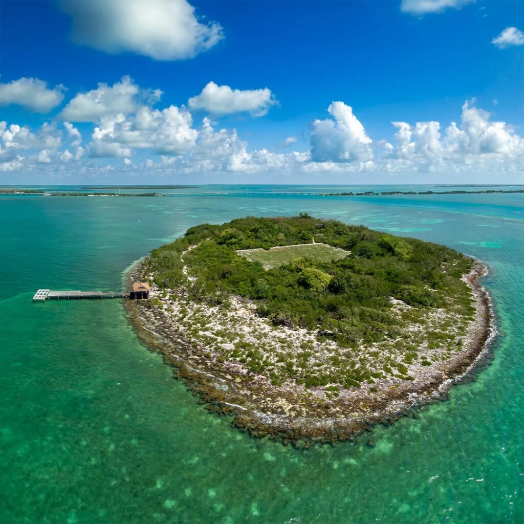 8 day trips from marathon fl you didn't know existed (2026) 5 A small, lush green island surrounded by clear turquoise water, with a dock extending from its shore. Puffy white clouds and a blue sky fill the background.
