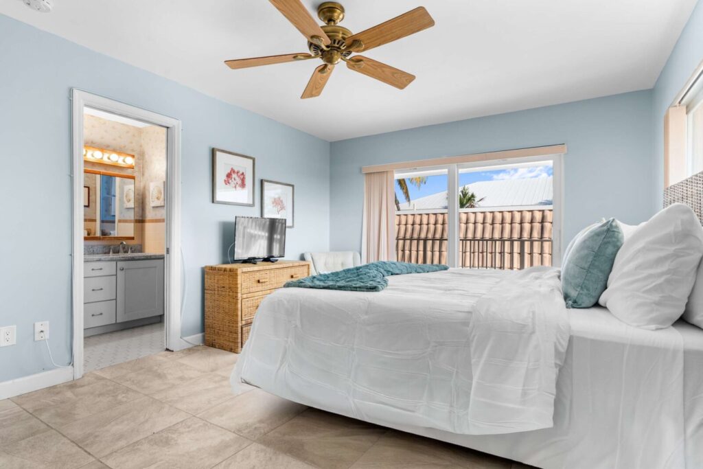 Bedroom with light blue walls, a white bed, wicker dresser with a tv, ceiling fan, large windows with a patio view, tile floors, and an open door leading to a bathroom with a large mirror and vanity lights.